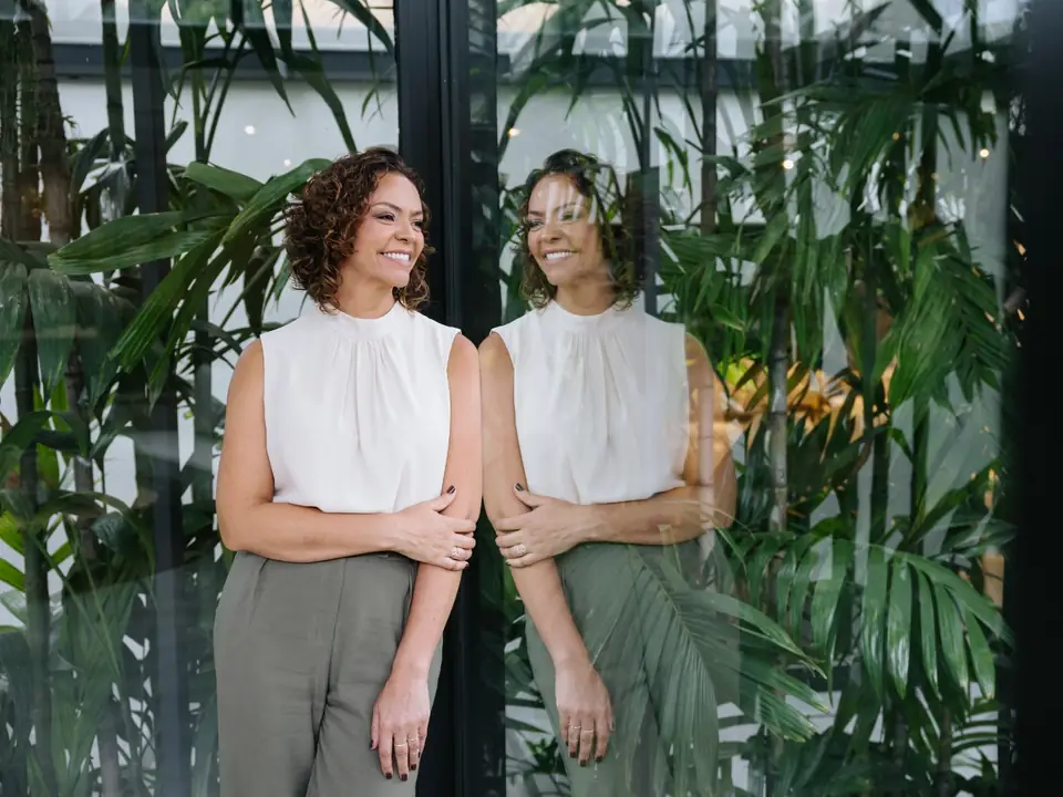 Débora Parreira de pé ao lado de uma parede de vidro, com o próprio reflexo e plantas ao fundo